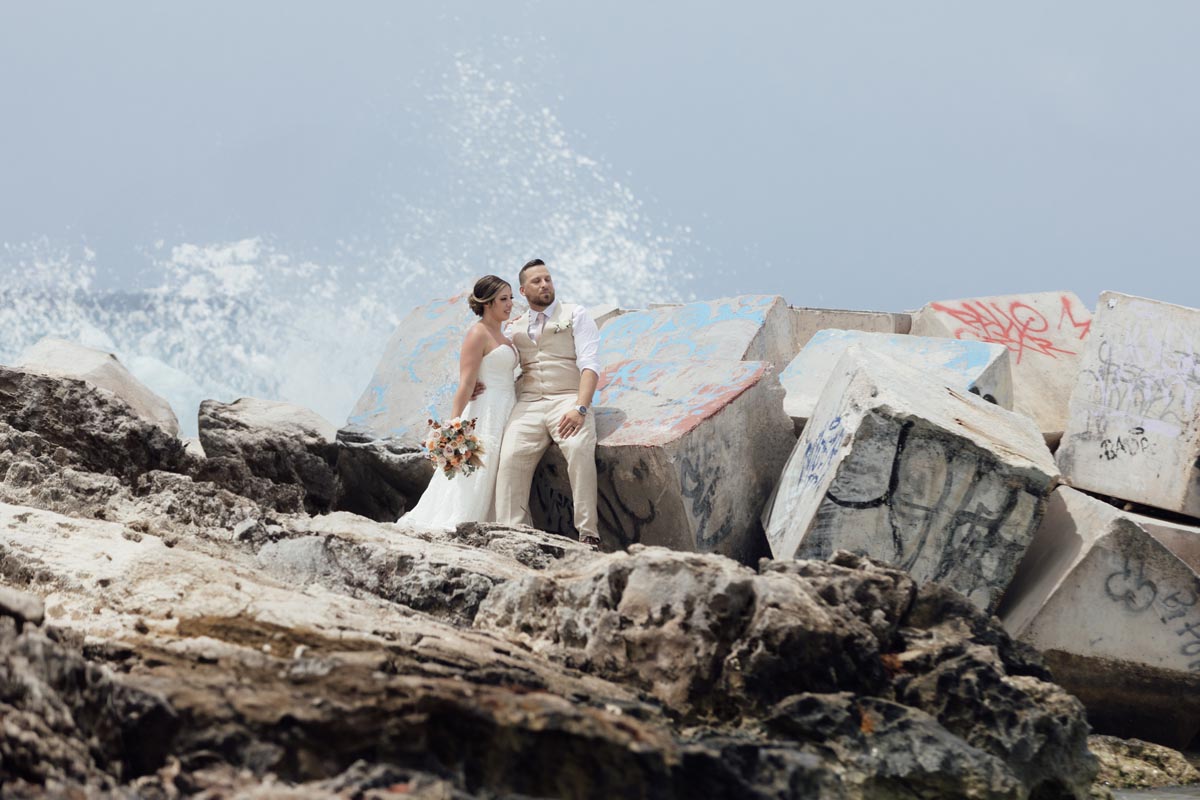 Newlyweds on rugged rocks with a giant ocean splash behind them