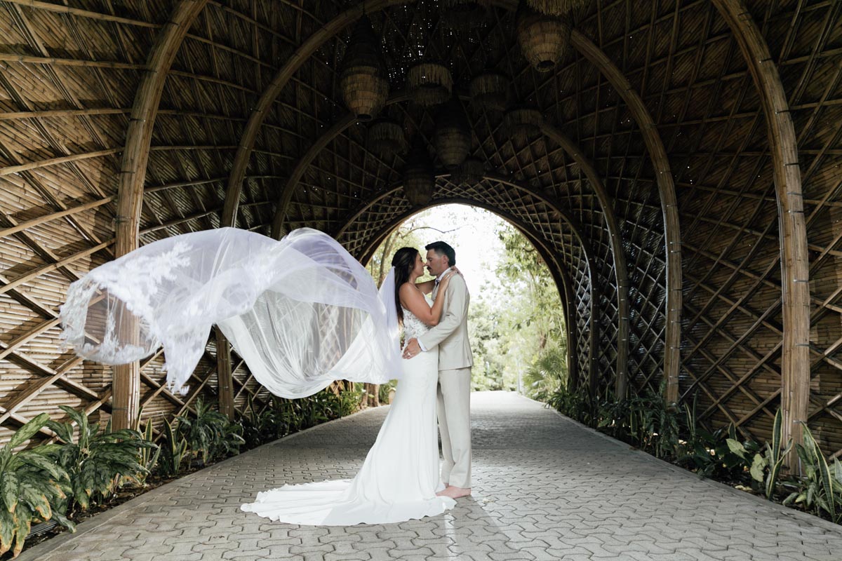 Newlyweds embrace in a bamboo tunnel with her veil floating on the breeze