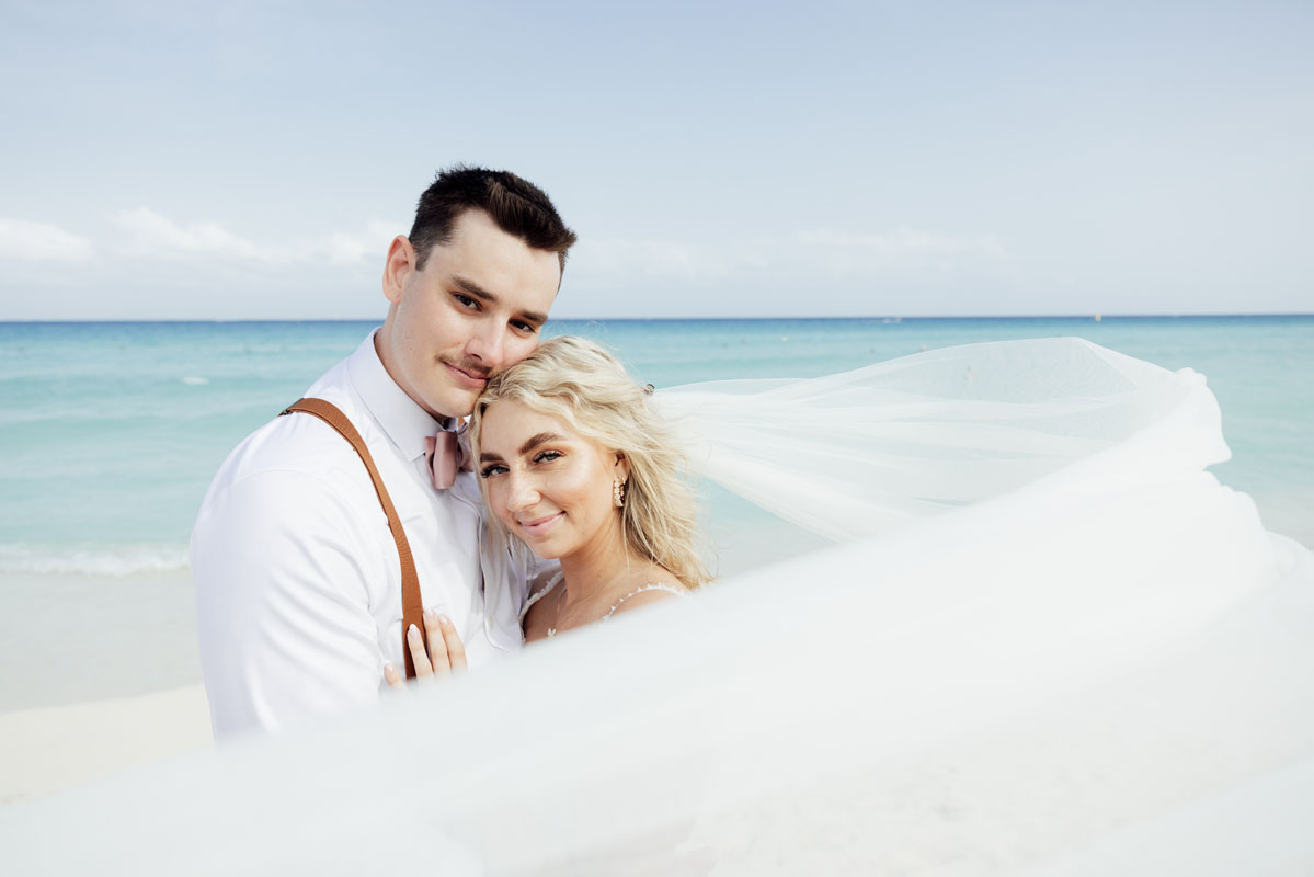 Glowing newlyweds embrace on beach behind brides billowing veil