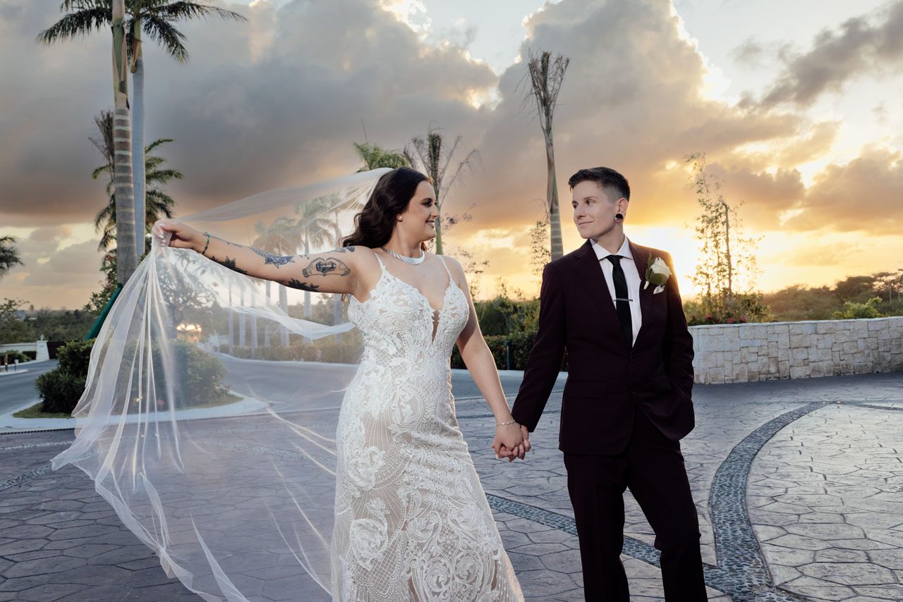 Newlyweds hold hands on resort street while bride billows veil in golden sunset