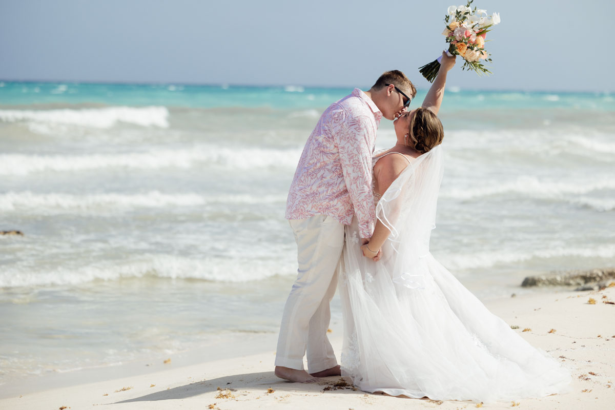 Newlyweds kiss on the beach as bride holds her bouquet up high