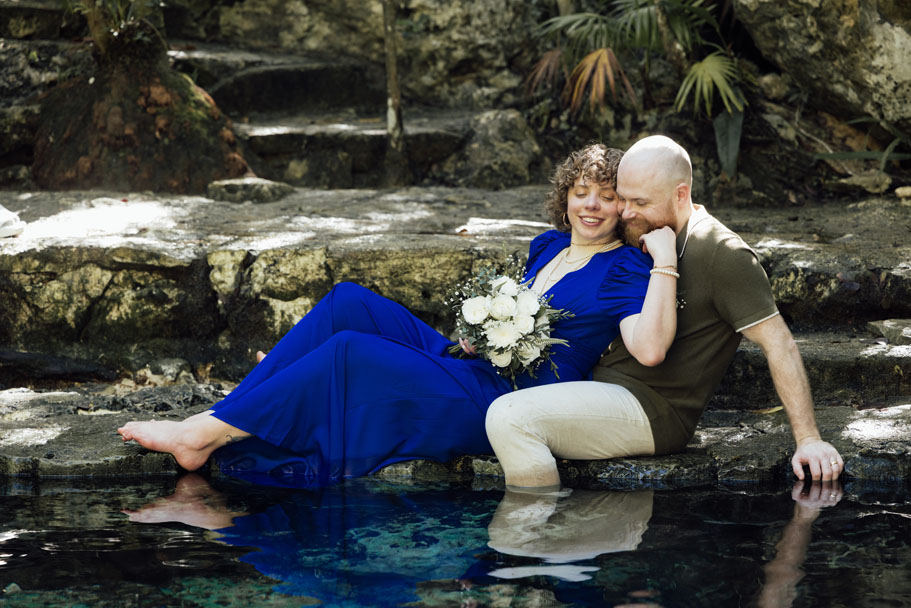 Smiling newlyeds embrace sitting at cenote water's edge